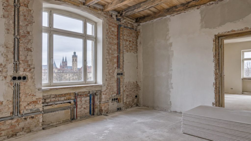 Altbauraum im Umbau mit freigelegtem Mauerwerk, Leitungen, Putzflächen und gestapelten Gipskartonplatten.